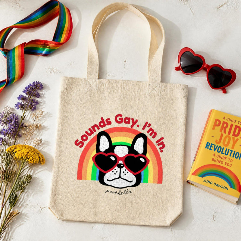 Tote bag with a dog graphic and rainbow text, surrounded by a book, sunglasses, and flowers on a light background