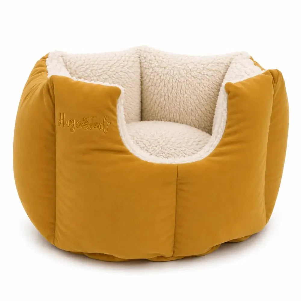 A round, low-profile pet bed with a mustard yellow velvet exterior and a cream sherpa fleece interior, featuring the brand name 'Hugo & Ted' embroidered on the left side.