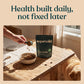 A person's hand pours kibble into a bowl next to a black stand-up pouch of superwild dog food on a rustic wooden table, with a golden retriever dog sitting nearby. The scene is framed with a beige border and the text 'Health built daily, not fixed later' at the top.