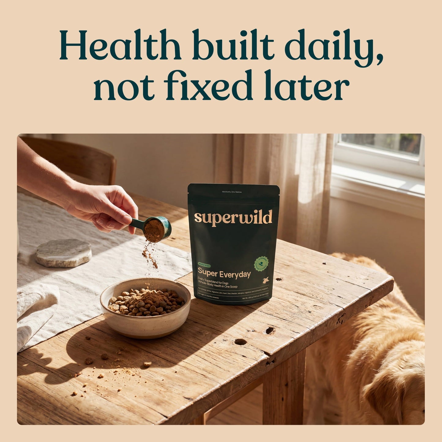A person's hand pours kibble into a bowl next to a black stand-up pouch of superwild dog food on a rustic wooden table, with a golden retriever dog sitting nearby. The scene is framed with a beige border and the text 'Health built daily, not fixed later' at the top.