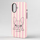 A phone case with vertical pink and cream stripes, featuring a black line art illustration of a French Bulldog peeking over the bottom edge with the word 'poochella' written in cursive below.