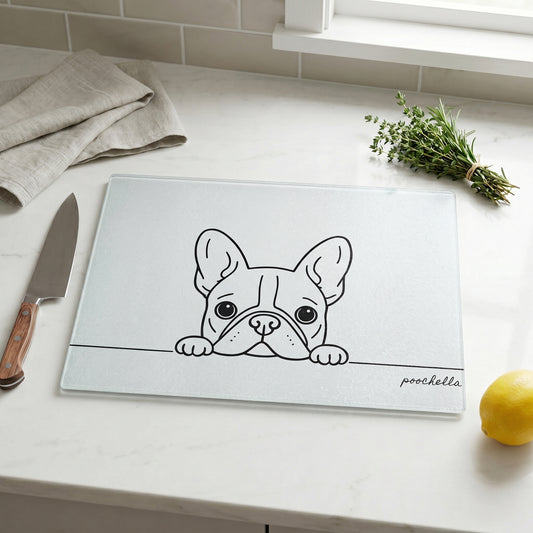 Glass cutting board with a french bulldog illustration on a kitchen counter with a knife, lemon, and herbs.