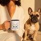 Person holding a mug with a dog design, sitting next to a french bulldog on a wooden floor.