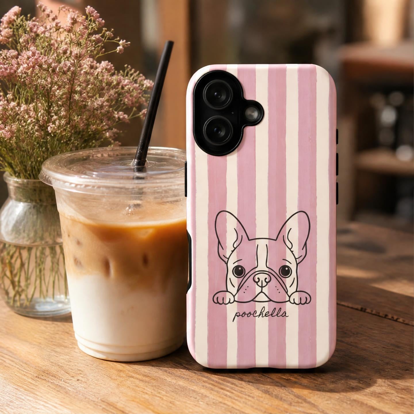 A pink and cream striped phone case with a cartoon French Bulldog illustration and the name 'poochella' printed on it, shown standing on a wooden table next to an iced coffee and a vase of flowers.