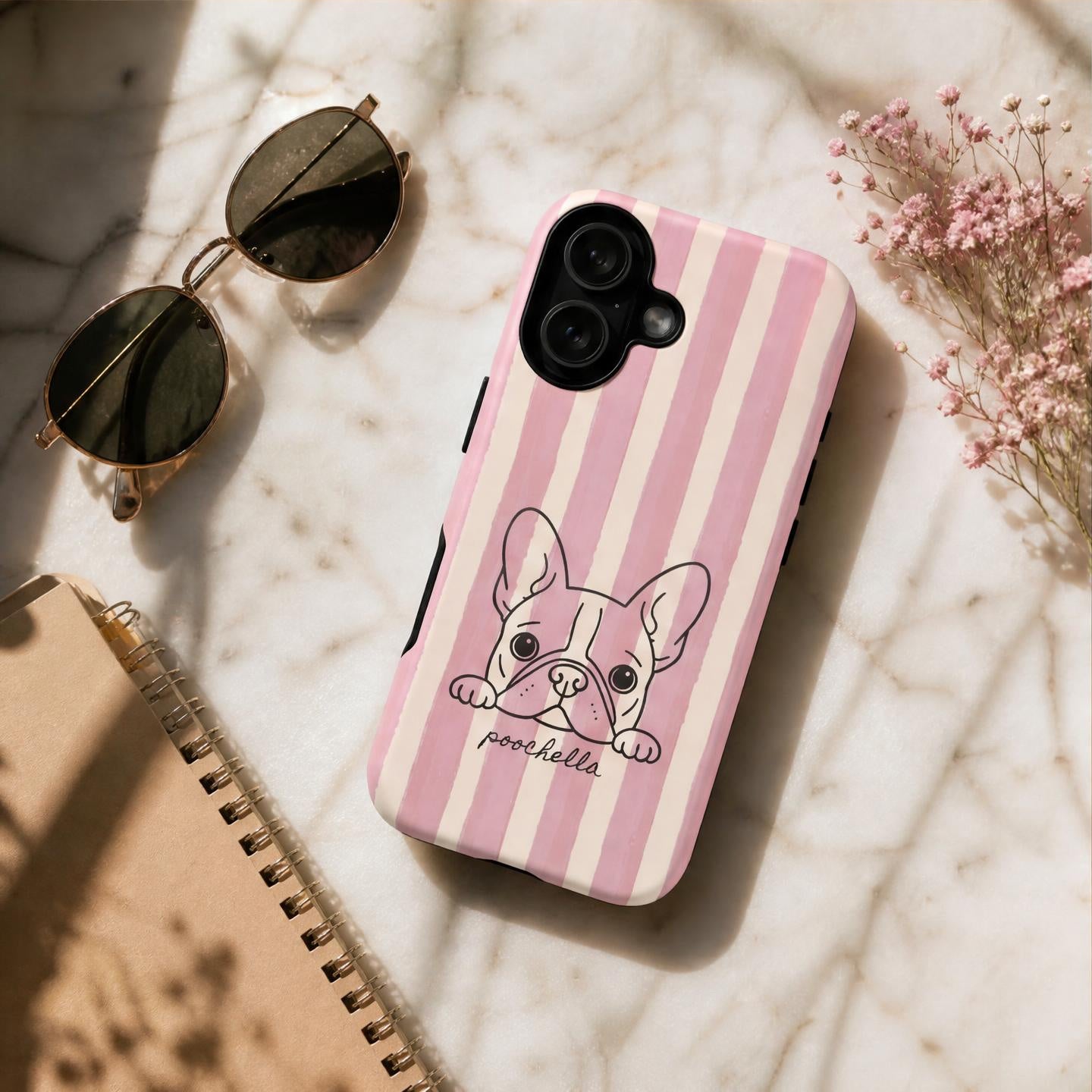 A pink and cream striped phone case with a cartoon French Bulldog illustration and the text 'poochella' lies on a white marble surface next to gold-framed sunglasses, a notebook, and small pink flowers.