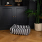 Black and white striped pet bed on a wooden floor with a dark cabinet and plant in the background.