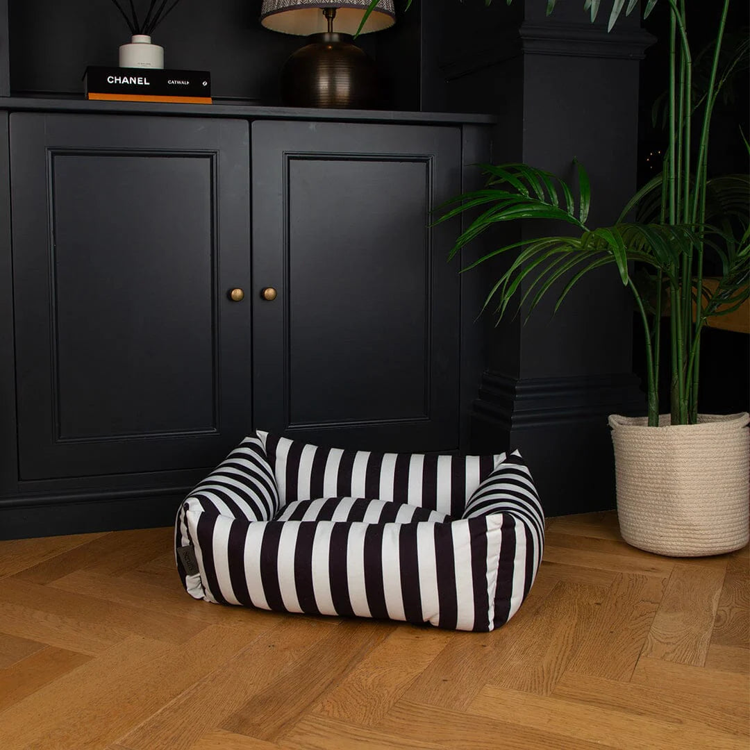 Black and white striped pet bed on a wooden floor with a dark cabinet and plant in the background.