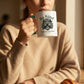 Person holding a mug with 'Be Kind Karma's Watching' text, sitting at a table with a croissant and newspaper.