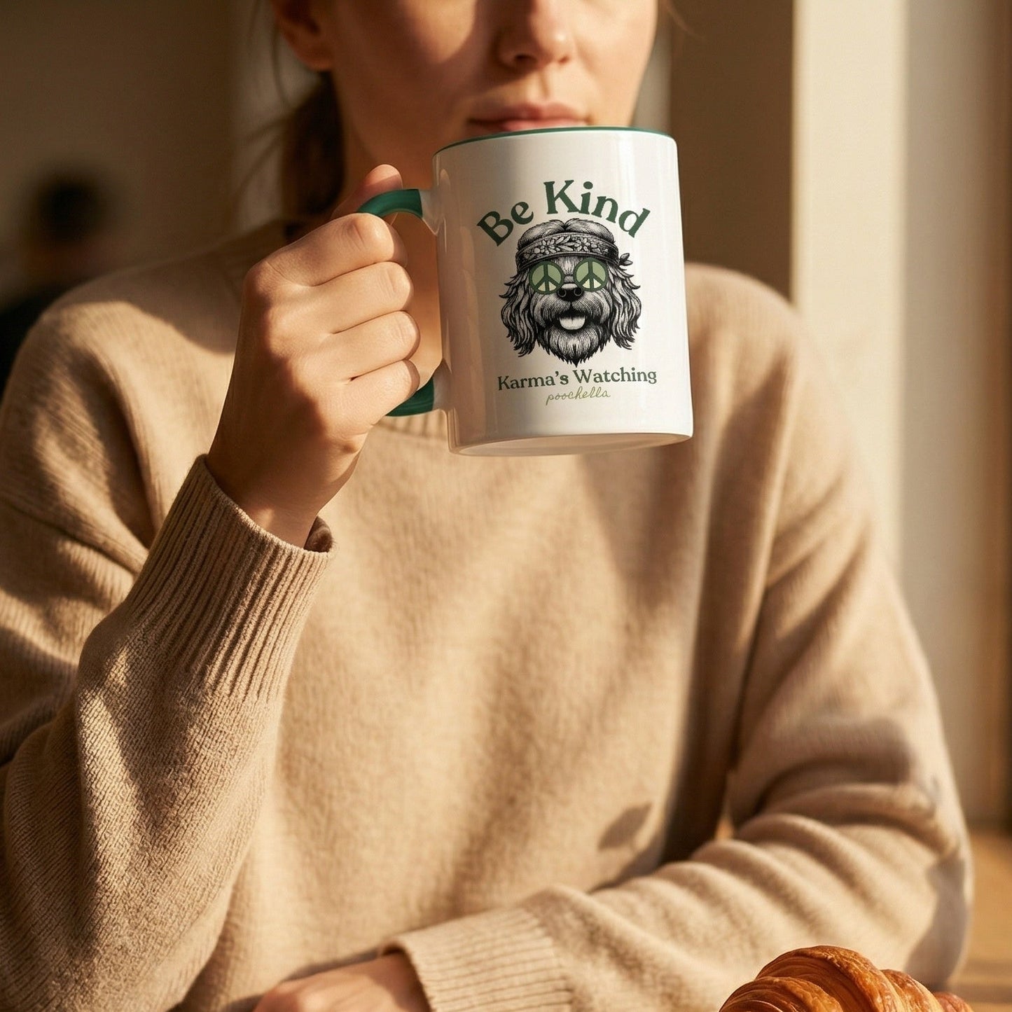 Person holding a mug with 'Be Kind Karma's Watching' text, sitting at a table with a croissant and newspaper.