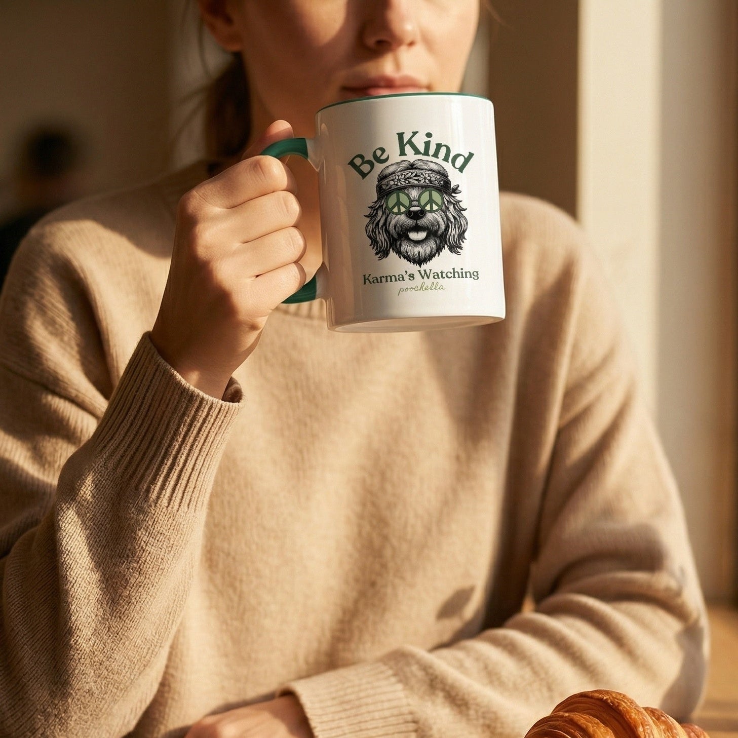 Person holding a mug with 'Be Kind Karma's Watching' text, sitting at a table with a croissant and newspaper.