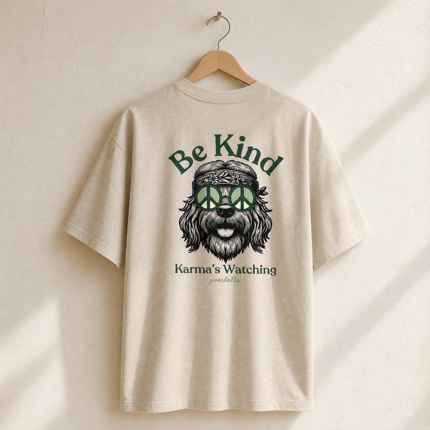 Beige t-shirt with a graphic of a dog wearing a peace sign headband and text 'Be Kind Karma's Watching' on a wooden hanger.