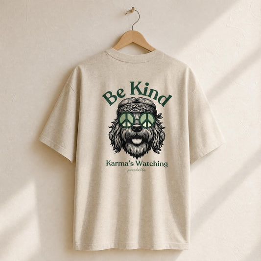 Beige t-shirt with a graphic of a dog wearing a peace sign headband and text 'Be Kind Karma's Watching' on a wooden hanger.