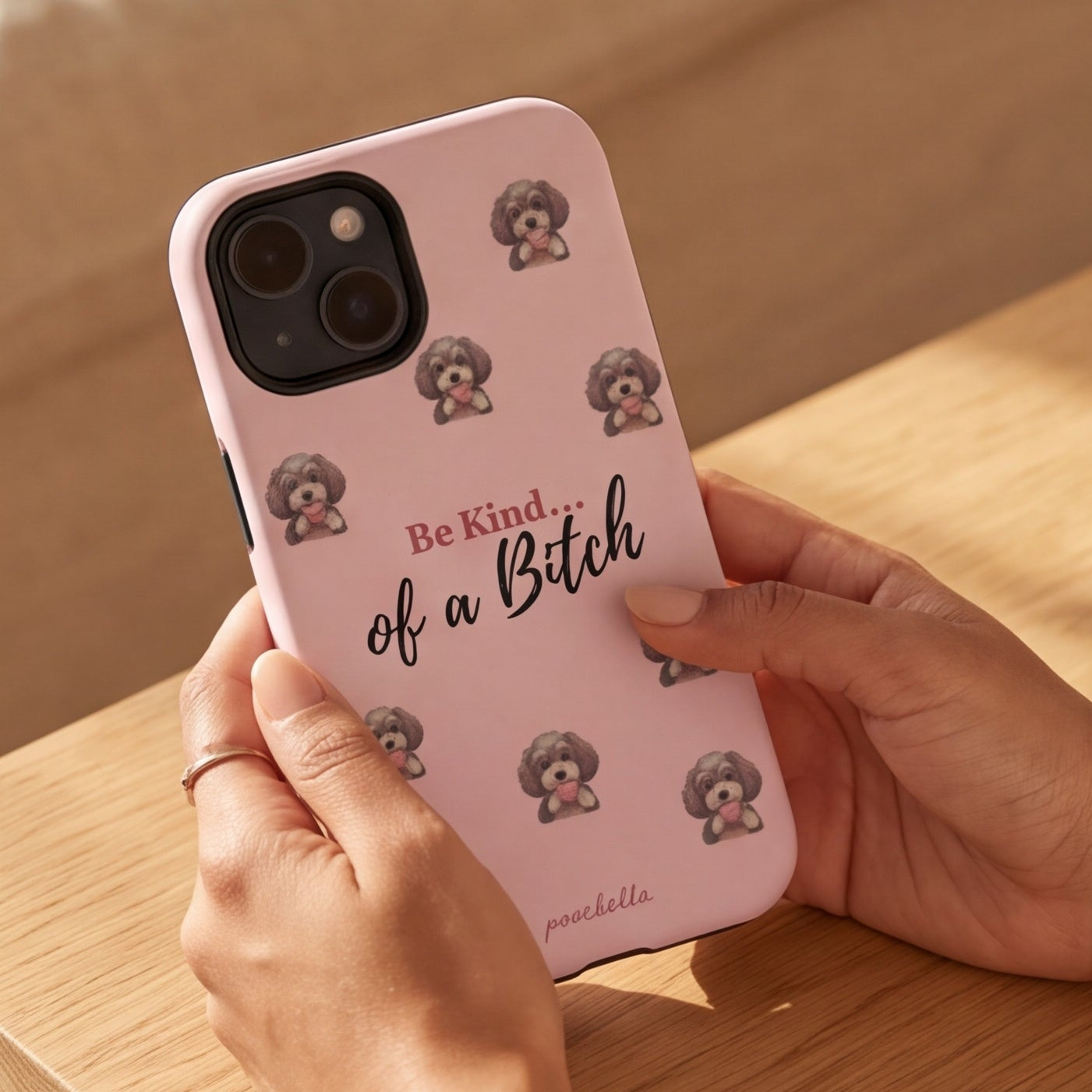 A person holds a light pink phone case featuring a cute cartoon dog print and the text 'Be Kind of a Bitch' in a warm, sunlit indoor setting.