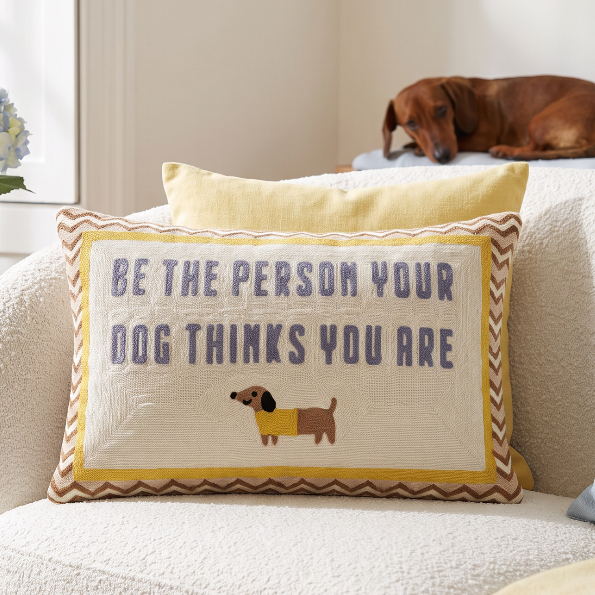A rectangular decorative throw pillow on a cream boucle sofa in a bright living room. The pillow has a cream center with the embroidered text 'BE THE PERSON YOUR DOG THINKS YOU ARE' in blue-gray, a small dachshund illustration in a yellow sweater, and a yellow border with brown chevron trim.