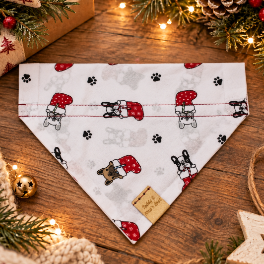 A white dog bandana with a Christmas-themed print of dogs in red Santa hats, paw prints, and snow globes, styled on a wooden table with holiday decorations.