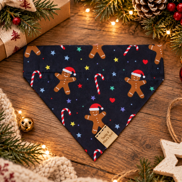 A navy blue bandana pet collar decorated with a Christmas gingerbread man and candy cane pattern, styled on a wooden tabletop with holiday decorations.