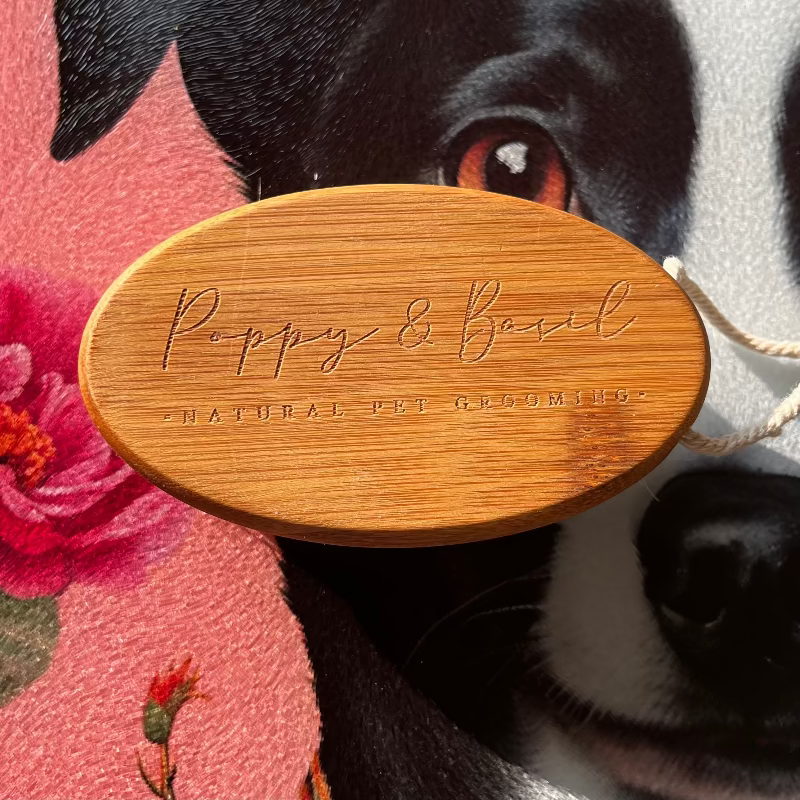 Wooden tag with 'Poppy & Basil' branding held by a dog in front of flowers