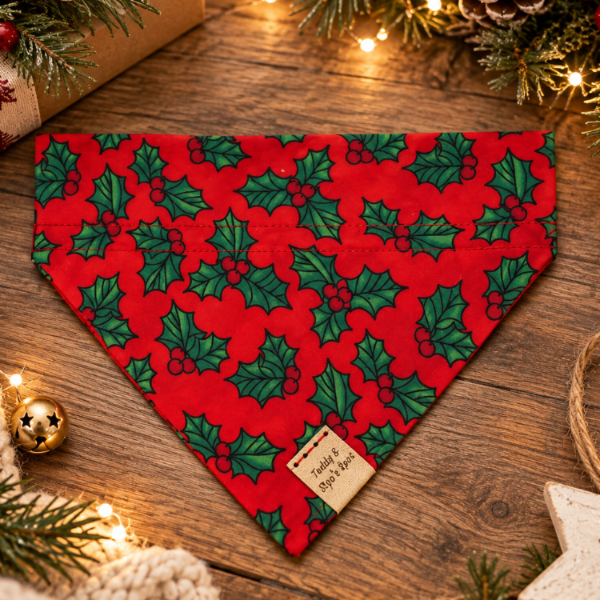 A red pet bandana with a green holly and red berry pattern, styled in a cozy Christmas flat-lay on a wooden surface with pine branches, fairy lights, a gift box, and a knitted blanket.