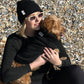 Person sitting on a pebbly beach holding a small brown dog dressed in a black dog jumper