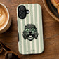 A phone case with a light green and white vertical stripe pattern, featuring a cartoon illustration of a shaggy dog wearing a bandana and peace-sign sunglasses. The case is displayed on a wooden table surrounded by lifestyle props, including a cup of coffee, sunglasses, and a croissant.