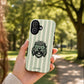 A hand holds a phone case featuring a green and white striped background with a black-and-white cartoon dog wearing a peace sign hat. The background is a blurred park scene with trees and a path.