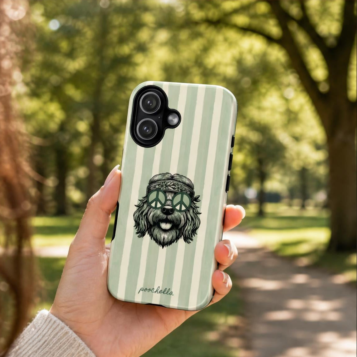 A hand holds a phone case featuring a green and white striped background with a black-and-white cartoon dog wearing a peace sign hat. The background is a blurred park scene with trees and a path.