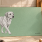 A rectangular green cutting board with a black-and-white sketch of a Labrador Retriever, displayed on a kitchen counter surrounded by bread, basil, and wooden utensils.