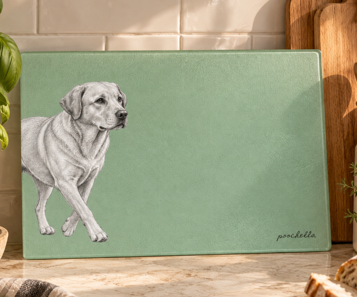 A rectangular green cutting board with a black-and-white sketch of a Labrador Retriever, displayed on a kitchen counter surrounded by bread, basil, and wooden utensils.