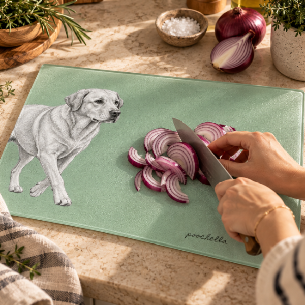 A mint green cutting board with a black-and-white illustration of a dog and the name 'poochella' in script, shown on a kitchen counter while a person slices a red onion with a knife.