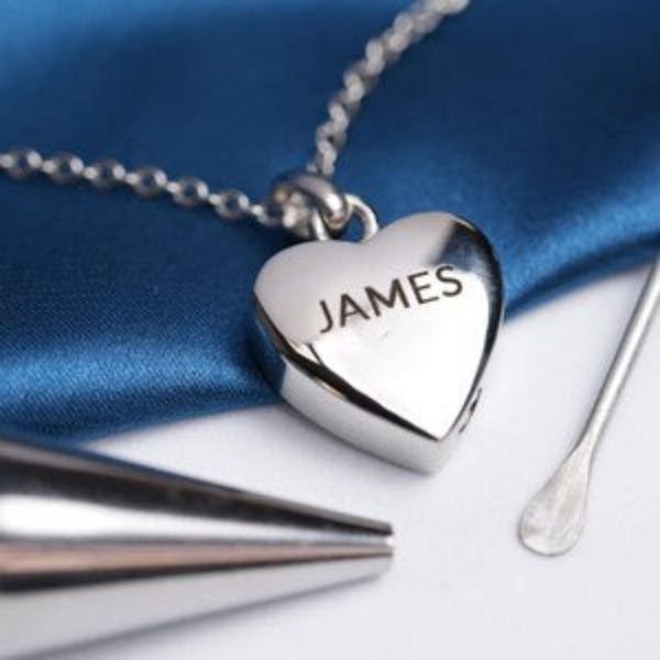 A silver heart-shaped pendant necklace engraved with the name 'JAMES', resting on a blue satin fabric background with metal tools nearby.