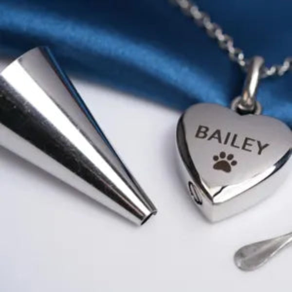 A silver heart-shaped pendant necklace engraved with the name 'BAILEY' and a paw print, shown alongside a silver cone-shaped tool and a small metal spatula on a white surface with a blue satin cloth in the background.
