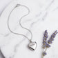 A silver heart pendant necklace engraved with the name 'JAMES' is displayed on a white marble plate next to dried lavender sprigs.