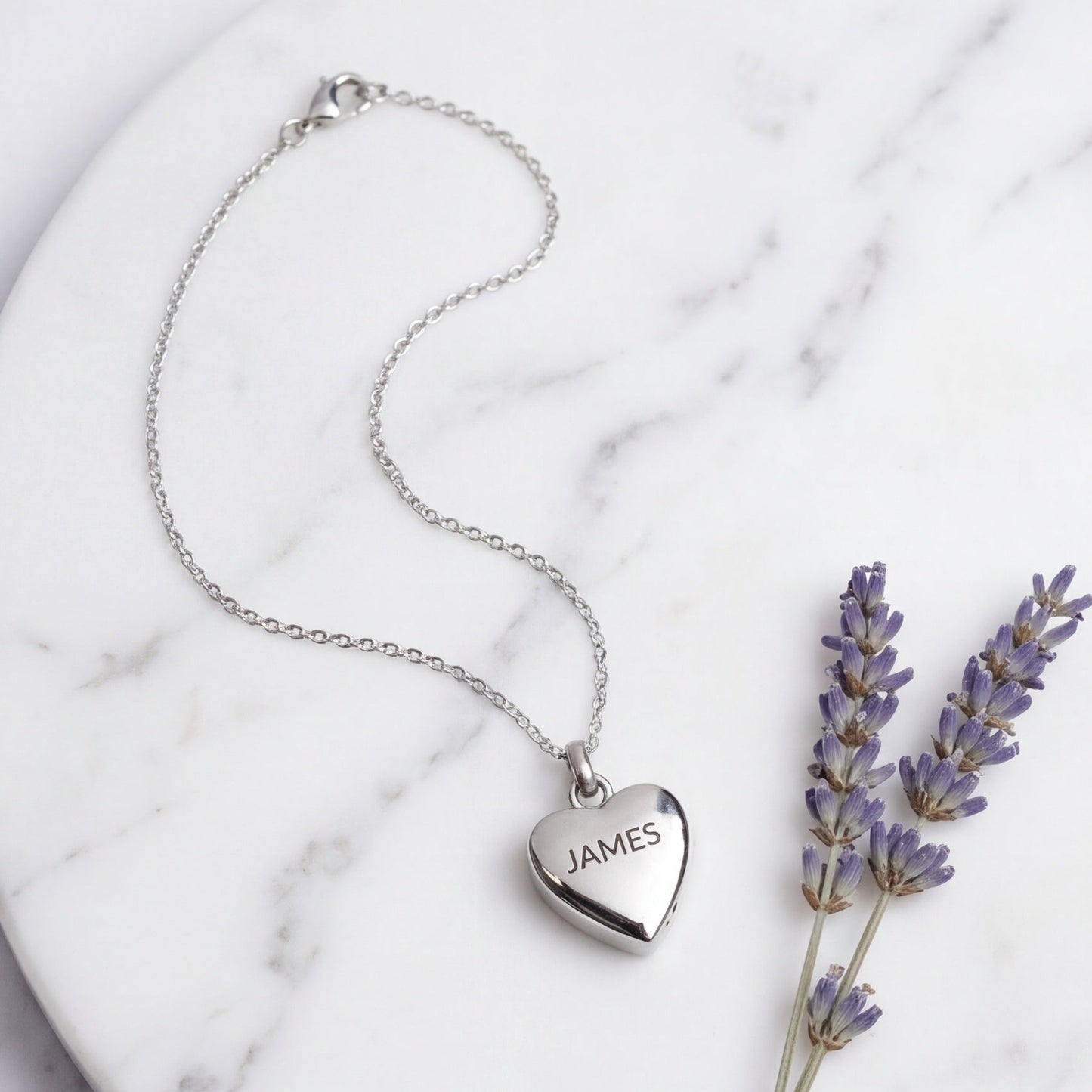 A silver heart pendant necklace engraved with the name 'JAMES' is displayed on a white marble plate next to dried lavender sprigs.