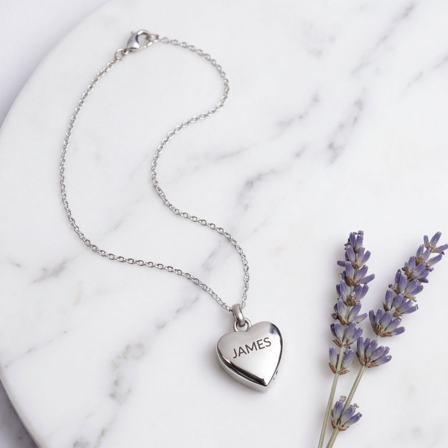 A silver heart pendant necklace engraved with the name 'JAMES' is displayed on a white marble plate next to dried lavender sprigs.