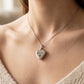 A close-up of a woman wearing a silver heart-shaped pendant necklace engraved with the name 'BAILEY' and a small paw print. The necklace is shown on a delicate silver chain against a warm, softly blurred indoor background.