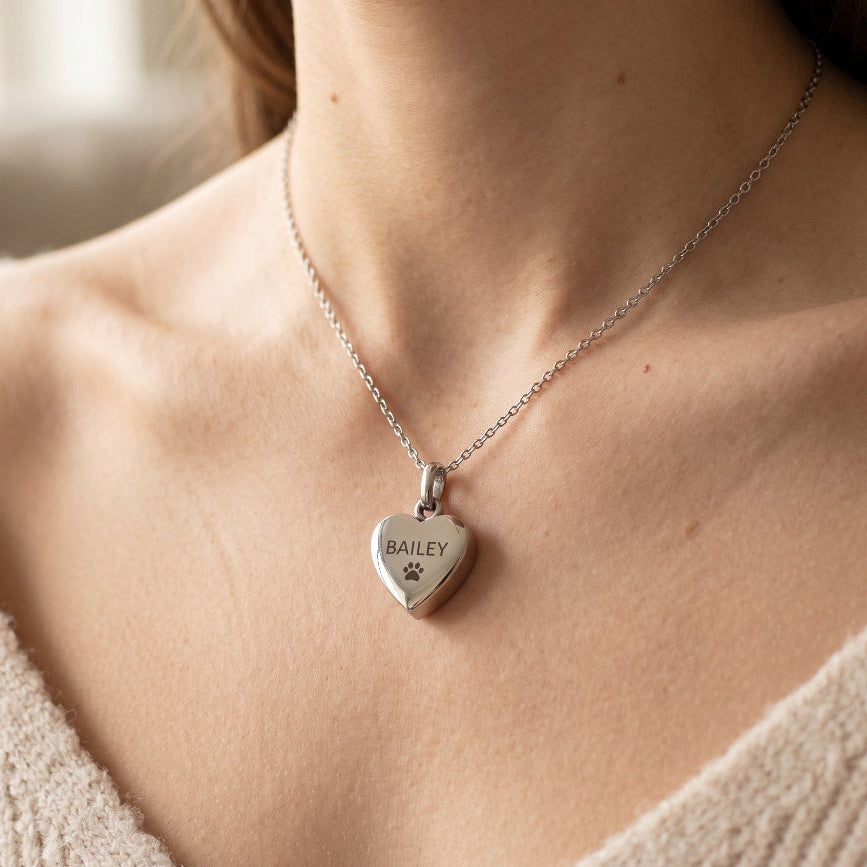 A close-up of a woman wearing a silver heart-shaped pendant necklace engraved with the name 'BAILEY' and a small paw print. The necklace is shown on a delicate silver chain against a warm, softly blurred indoor background.