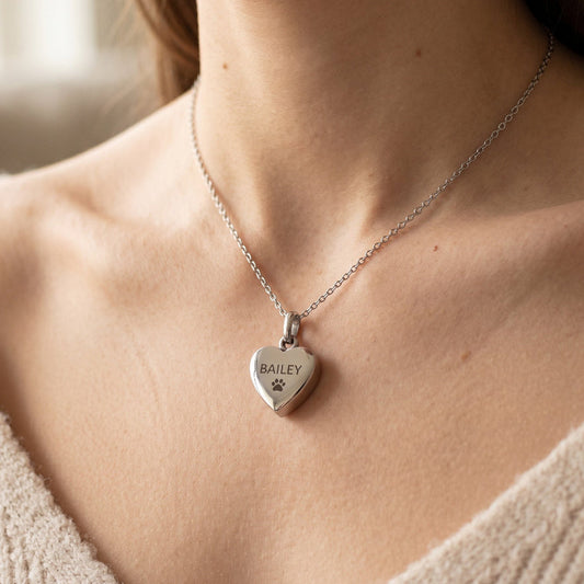 A close-up of a woman wearing a silver heart-shaped pendant necklace engraved with the name 'BAILEY' and a small paw print. The necklace is shown on a delicate silver chain against a warm, softly blurred indoor background.