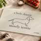 A rectangular glass cutting board with a frosted cream surface, printed with a black line drawing of a dachshund and the text 'a little chaotic mostly iconic' by poochella. The board is styled on a marble countertop with kitchen props including tomatoes, basil, a towel, and a bottle of olive oil.