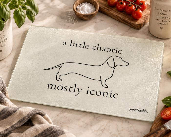 A rectangular glass cutting board with a frosted cream surface, printed with a black line drawing of a dachshund and the text 'a little chaotic mostly iconic' by poochella. The board is styled on a marble countertop with kitchen props including tomatoes, basil, a towel, and a bottle of olive oil.