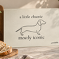 A rectangular ivory cutting board with a minimalist black line drawing of a dachshund and the text 'a little chaotic mostly iconic' by poochella, styled on a marble countertop with bread, basil, tomatoes, and kitchen utensils.