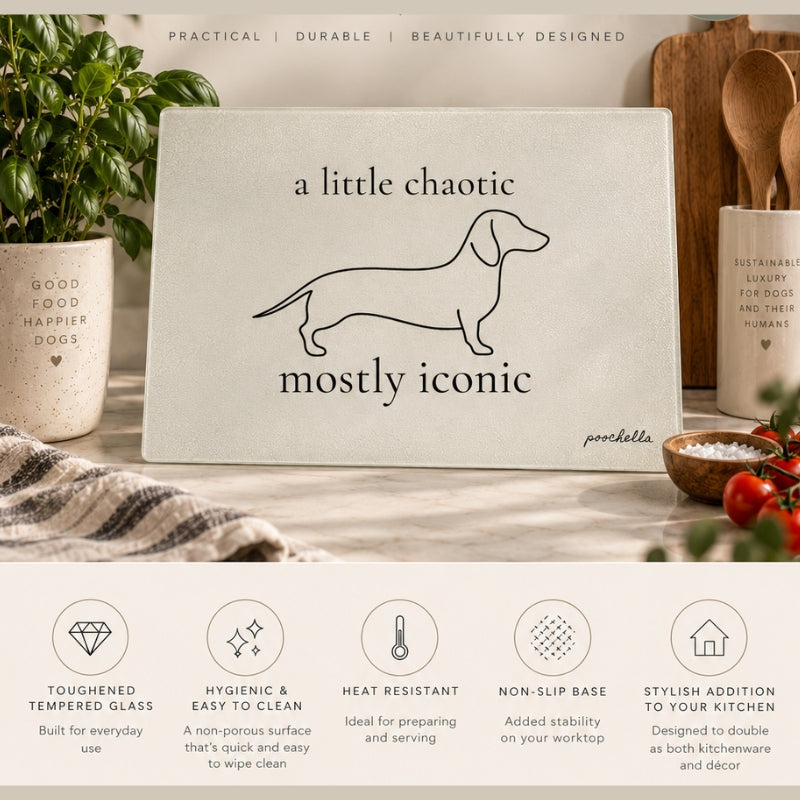 A rectangular kitchen cutting board with rounded corners, featuring a minimalist black line art illustration of a dachshund and the text 'a little chaotic mostly iconic' by the brand poochella. The board is styled on a wooden countertop with kitchen props like a potted plant, a candle, and tomatoes, with feature callout icons displayed along the bottom.