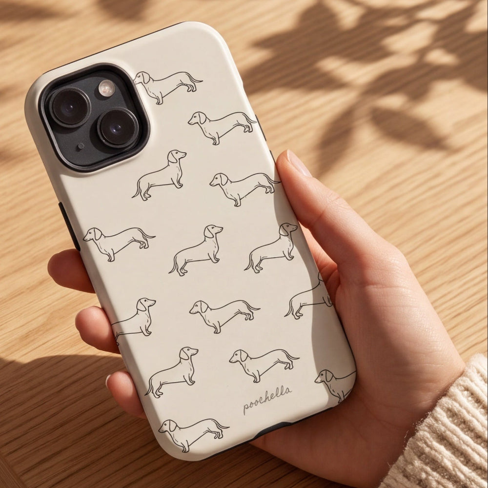 A hand holding a cream-colored phone case decorated with a repeating pattern of black line drawings of dachshunds, set against a warm wooden table background with leaf shadows.