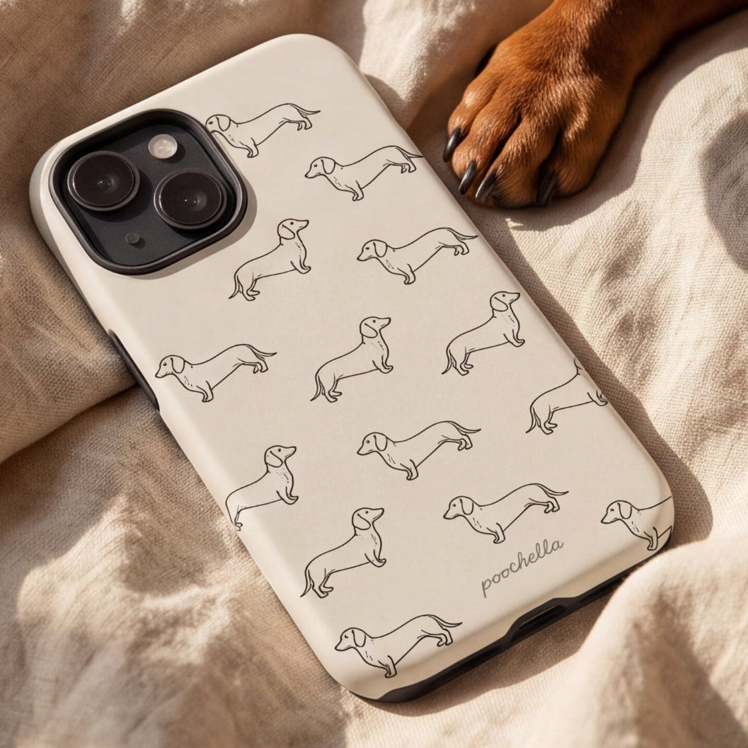 A cream-colored phone case with a black outline print of dachshunds, shown on a phone and placed on a beige linen blanket next to a brown dog's paw.