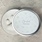 Round silver container with 'Cherished Keepsake' text on a textured surface