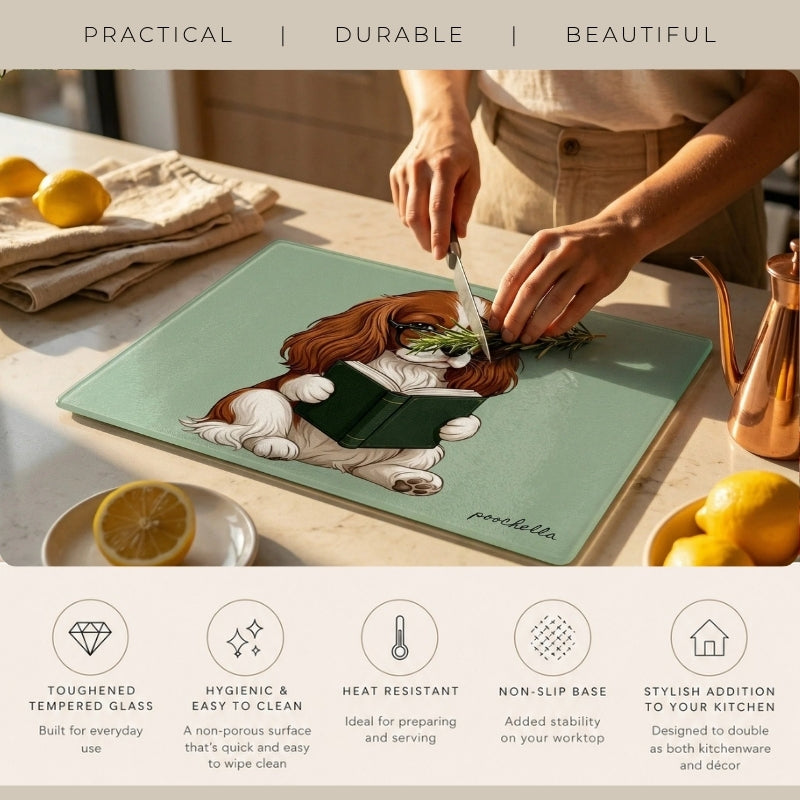 A kitchen scene showing a person slicing an avocado on a square mint green glass cutting board with a printed illustration of a dog reading a book. The board is placed on a countertop with lemons and kitchen items nearby.