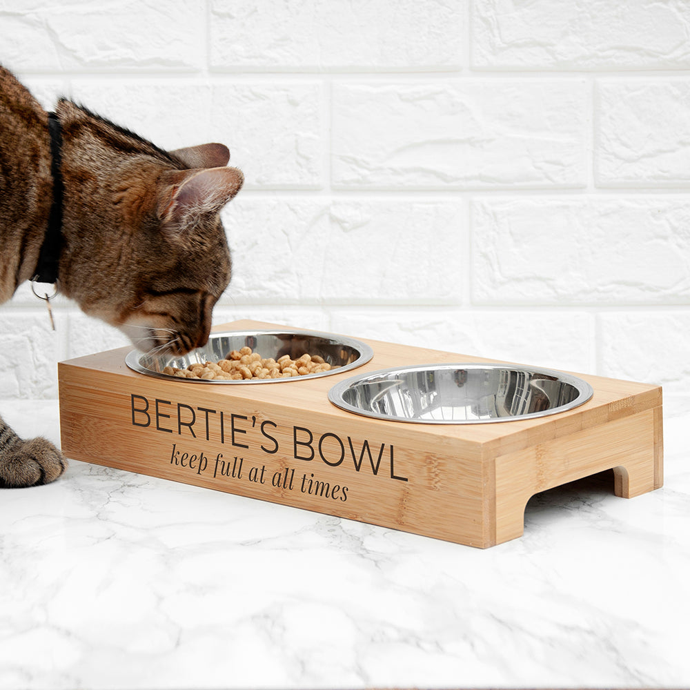Cat eating from a wooden pet bowl named 'Bertie's Bowl' on a white marble surface.