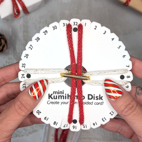 Kumihimo disk with red braided cord held by hands, with Christmas decorations in the background.