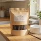 A kraft paper stand-up pouch labeled 'Poochella Natural Dog Chews Beef Pizzle Sticks' stands on a wooden kitchen counter with a white bowl nearby and a softly blurred kitchen background.
