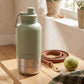 Green insulated bottle on a wooden table with a plaid towel and dog accessories.