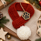 A red crocheted pet scarf with a white pom-pom and a paw-print wooden tag, styled on a linen surface with Christmas greenery, lights, a pinecone, gold ornaments, and a wrapped gift box.